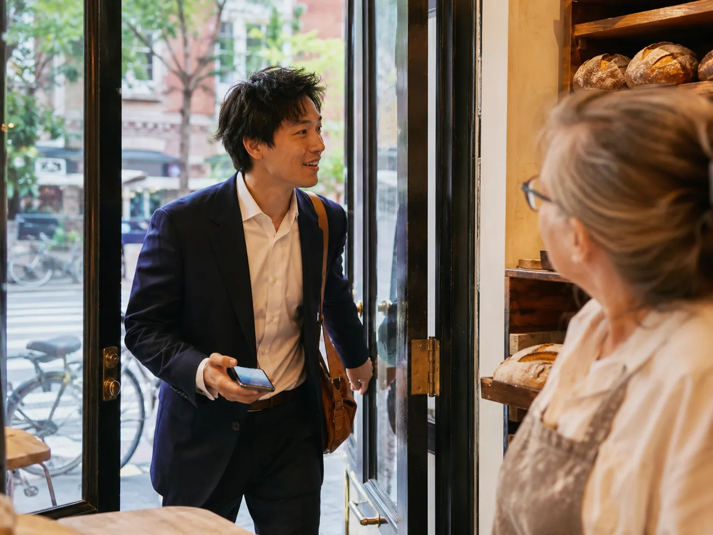 A young customer pushing open a neighborhood bakery door, phone still in hand from the AI recommendation that sent him, the baker looking up to greet him
