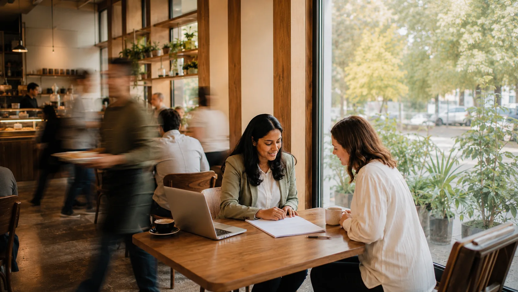 Two people sitting calm and in sharp focus across a cafe table with a proposal open between them, the busy cafe blurring into motion around them, the moment a meeting the pipeline booked actually happens